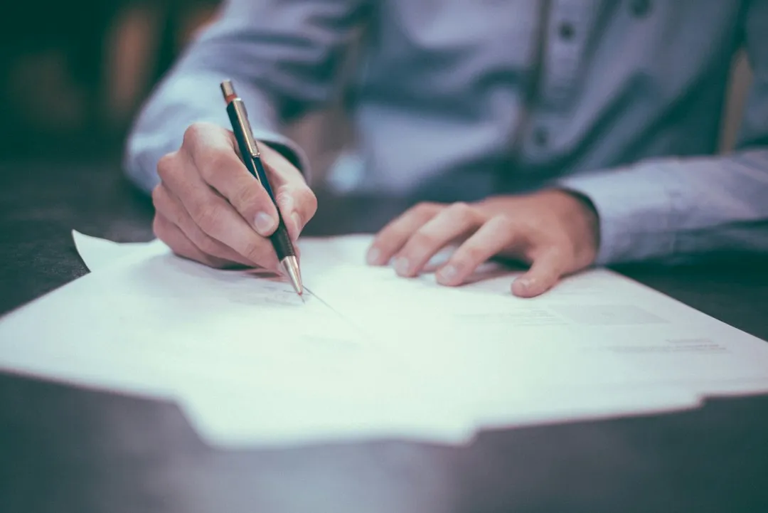 Main d'un homme en chemise qui signe un document papier au stylo, sur un bureau clair