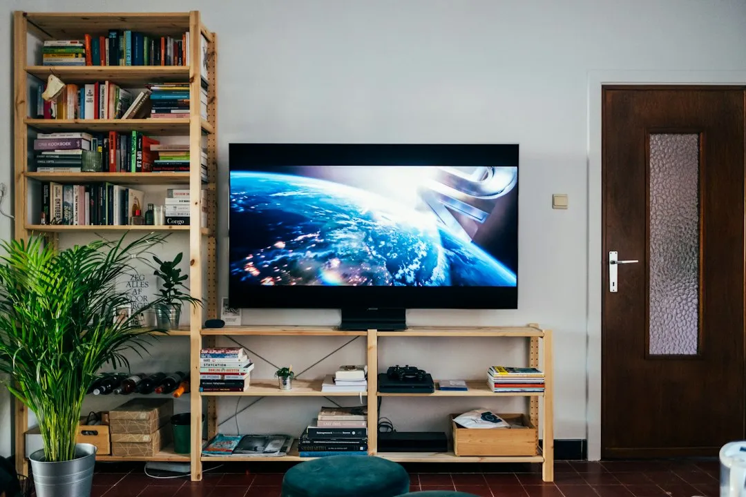 Téléviseur à écran plat allumé posé sur une étagère en bois dans un salon, livres et plantes autour