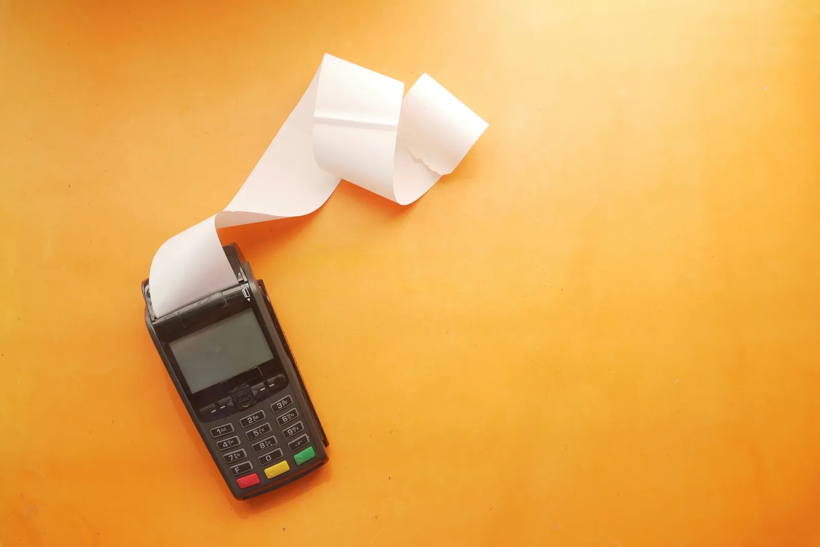 Terminal de paiement noir avec un long ticket de caisse en papier blanc, posé sur un fond orange