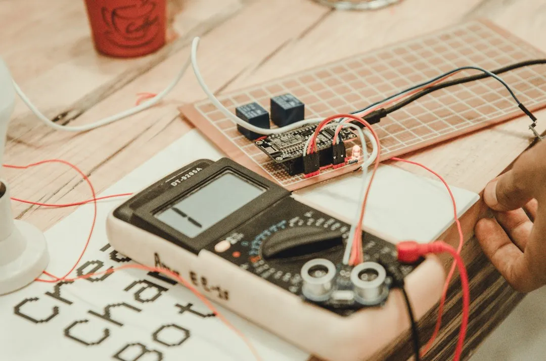 Mains d'un technicien qui mesurent un circuit électronique avec un multimètre blanc posé sur l'établi